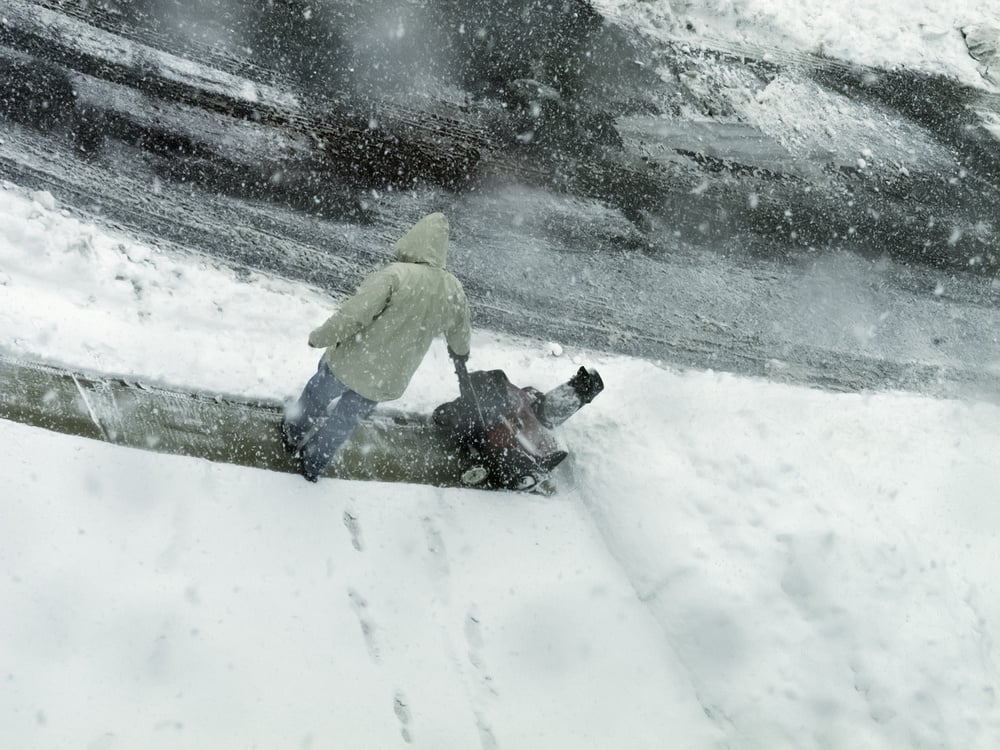 Winter at a glance Unrecognizable man in hooded coat stands holding a snow blower while he looks downward and snow falls all around him